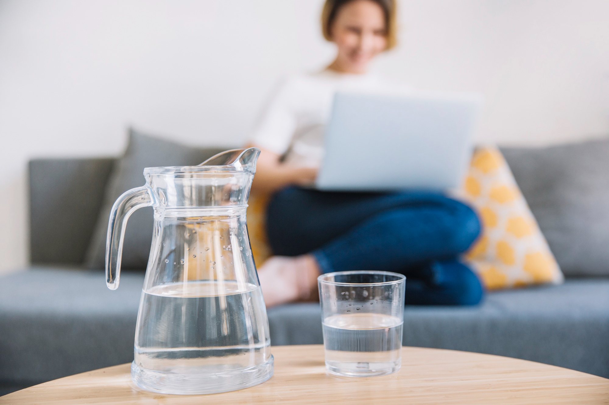 A jug and a glass of water on the foreground while a working woman on a laptop sits on a sofa in the background